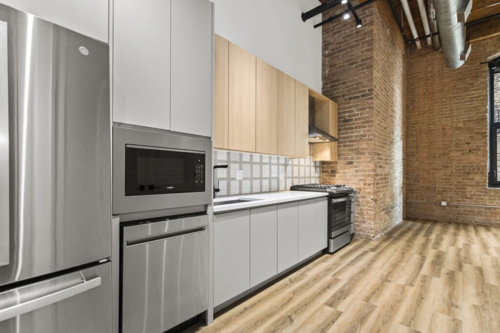 Modern kitchen with stainless steel appliances, light wood cabinets, white countertops, tile backsplash, exposed brick walls, wood flooring, and visible ceiling ductwork.