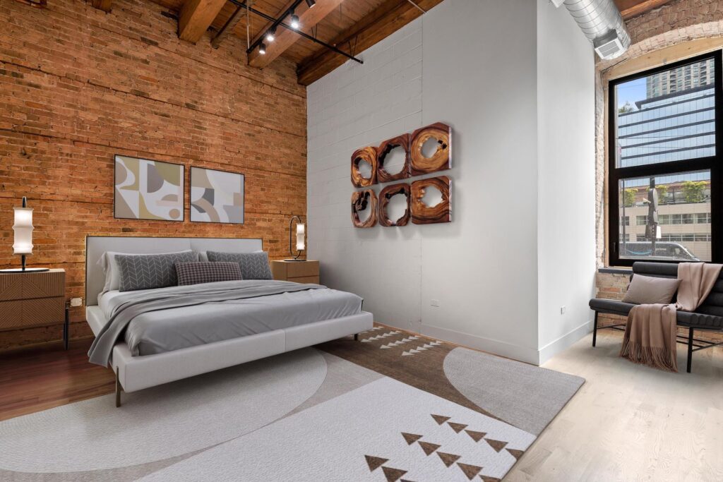 Modern bedroom with exposed brick wall, wooden ceiling beams, a large bed, abstract art, a window letting in natural light, and a chair in the corner.