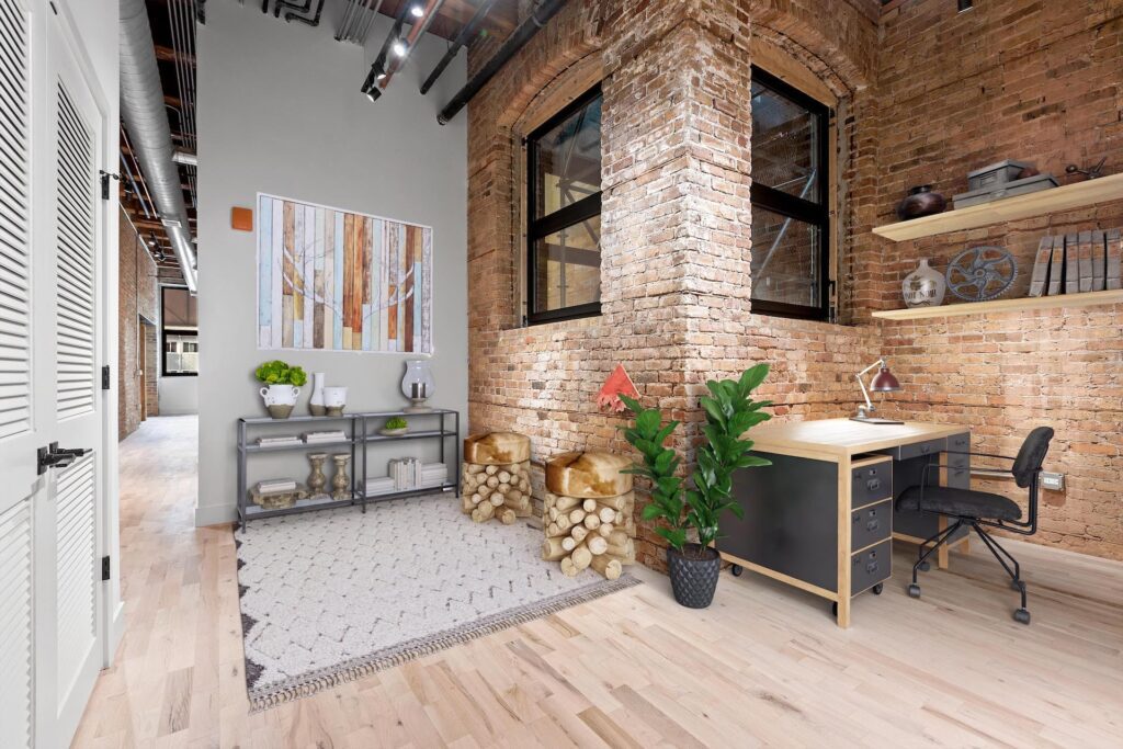 Modern office space with exposed brick walls, a wooden desk, shelves with books and décor, indoor plants, stacked firewood, and wall art above a side table.