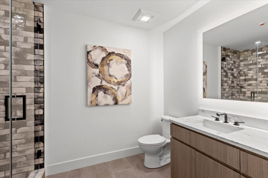 Modern bathroom with a glass-enclosed shower, abstract wall art above the toilet, a large mirror, and a wood vanity with a marble countertop.