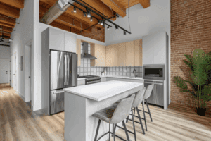 Modern kitchen with light wood cabinets, stainless steel appliances, white island with three gray chairs, exposed brick wall, and a potted plant.