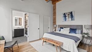 A modern bedroom with a neatly made bed, a wooden bench at the foot, two bedside tables with lamps, and an open door leading to a bathroom with a dark vanity.