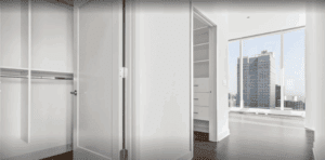 A modern, empty walk-in closet with built-in shelves and a partially open door, leading to a bright room with large windows and a city view.