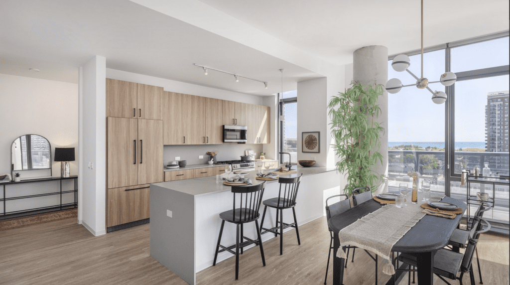 Modern apartment kitchen and dining area with light wood cabinets, black chairs, large windows, city views, and a potted plant by the dining table.