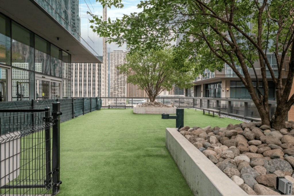 Outdoor terrace with artificial grass, a fenced area, trees, large planters with rocks, and city buildings visible in the background.