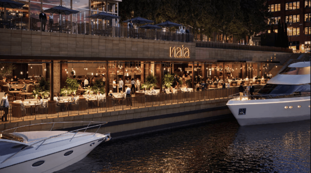 Two yachts are docked by a waterfront restaurant named "naia," with outdoor seating areas and people dining in the evening.