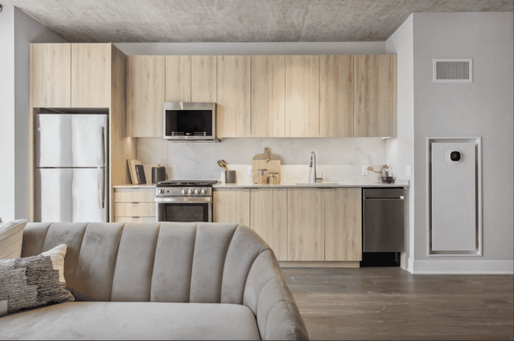 Modern kitchen with light wood cabinets, stainless steel appliances, and a gray sofa in the foreground. White countertop and backsplash, with minimal decor on the counters.