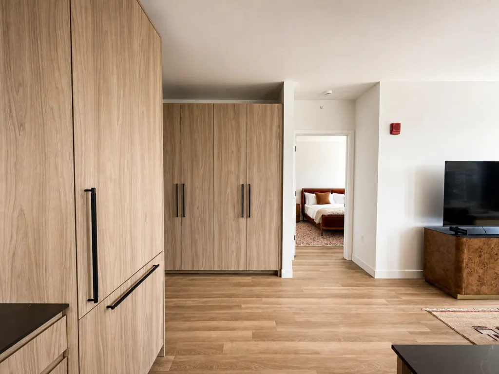 Modern apartment interior with wood-paneled cabinets, TV on a stand, and a view into a bedroom with a made bed. Light wood floors and white walls throughout.