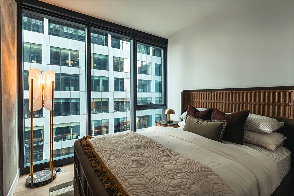 Modern bedroom with a large bed, textured headboard, floor lamp, gold desk lamp, and floor-to-ceiling windows overlooking a contemporary office building.