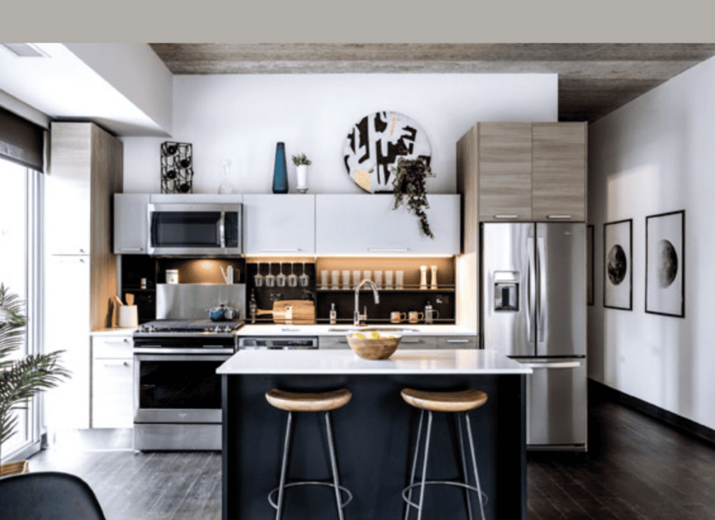 Modern kitchen with a central island, two wooden stools, stainless steel appliances, light wood cabinets, and decorative accents on shelves and walls.