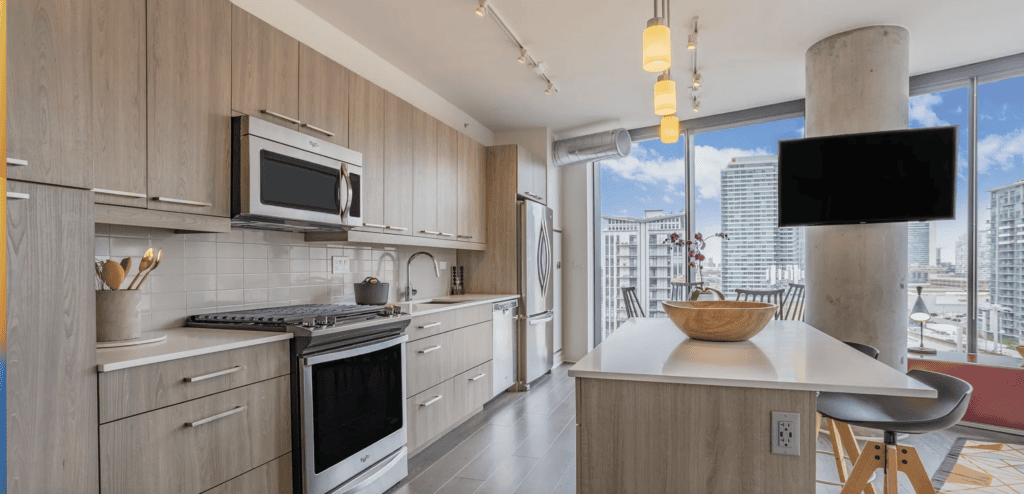 Modern kitchen with light wood cabinets, stainless steel appliances, island with sink, and large windows showing city views. Pendant and track lighting illuminate the space.