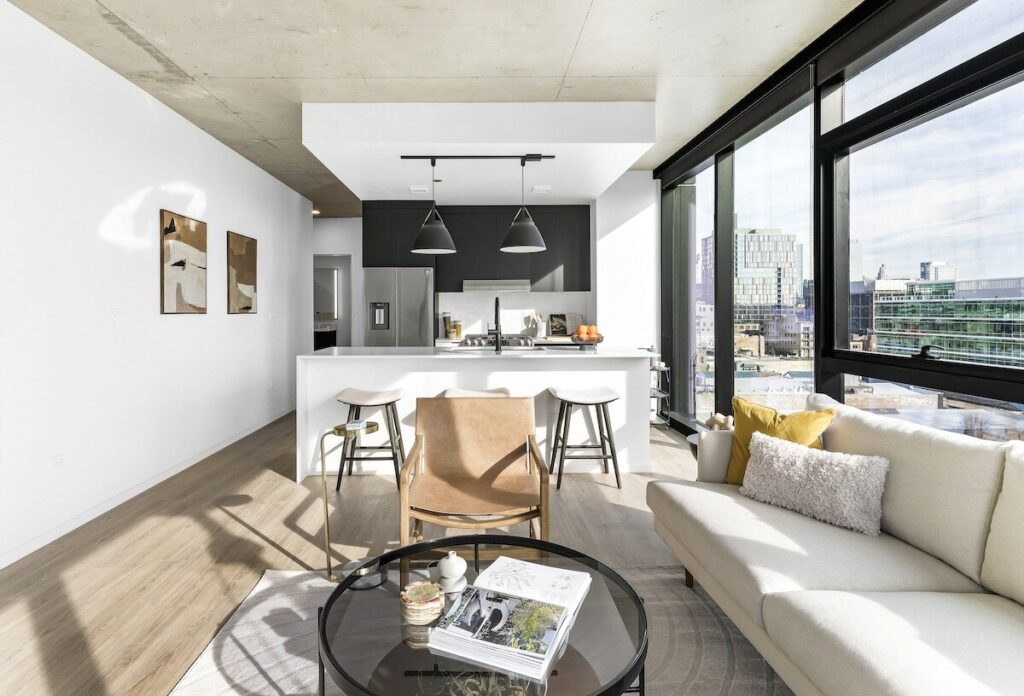 Luxury apartments at The Elizabeth by Sterling Bay. Modern open-plan living room with a kitchen island, two bar stools, a leather armchair, a glass coffee table, and a white sofa, with floor-to-ceiling windows overlooking a city skyline.