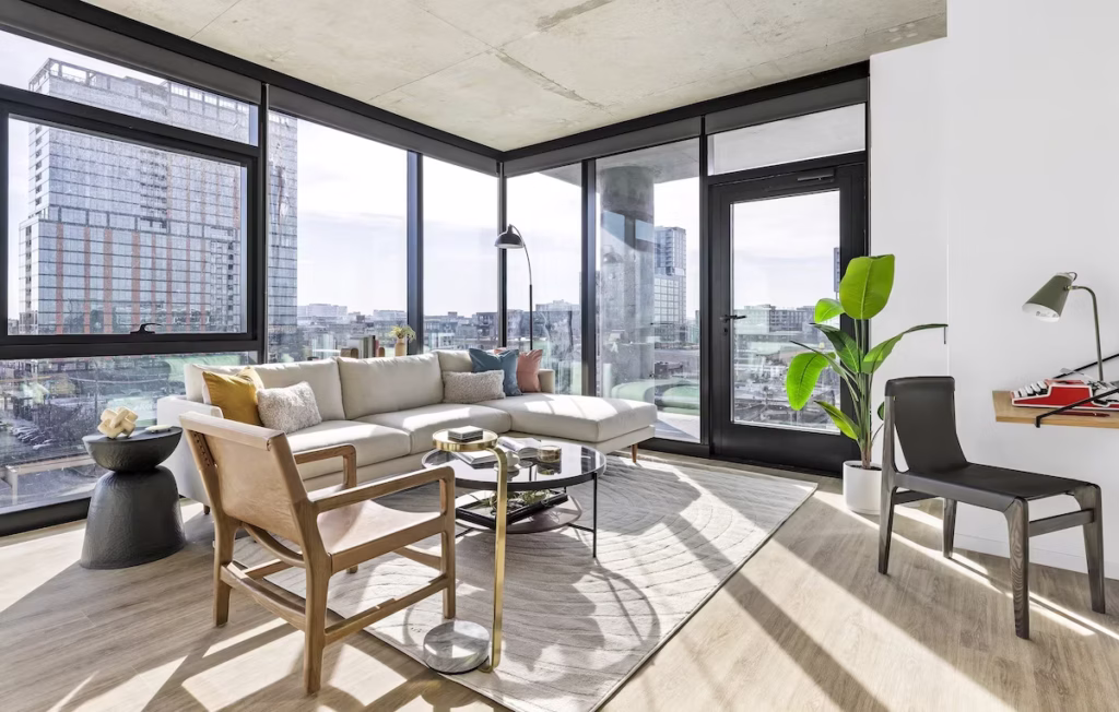 Luxury apartments at The Elizabeth by Sterling Bay. A modern living room with floor-to-ceiling corner windows, a sectional sofa, a wooden armchair, and a small desk area with a typewriter.