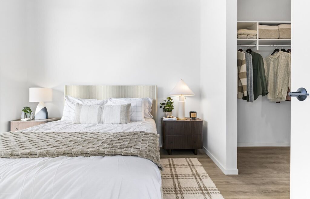 Luxury apartments at The Elizabeth by Sterling Bay. A bedroom with a queen bed, two nightstands with lamps, and an open walk-in closet with hanging clothes.