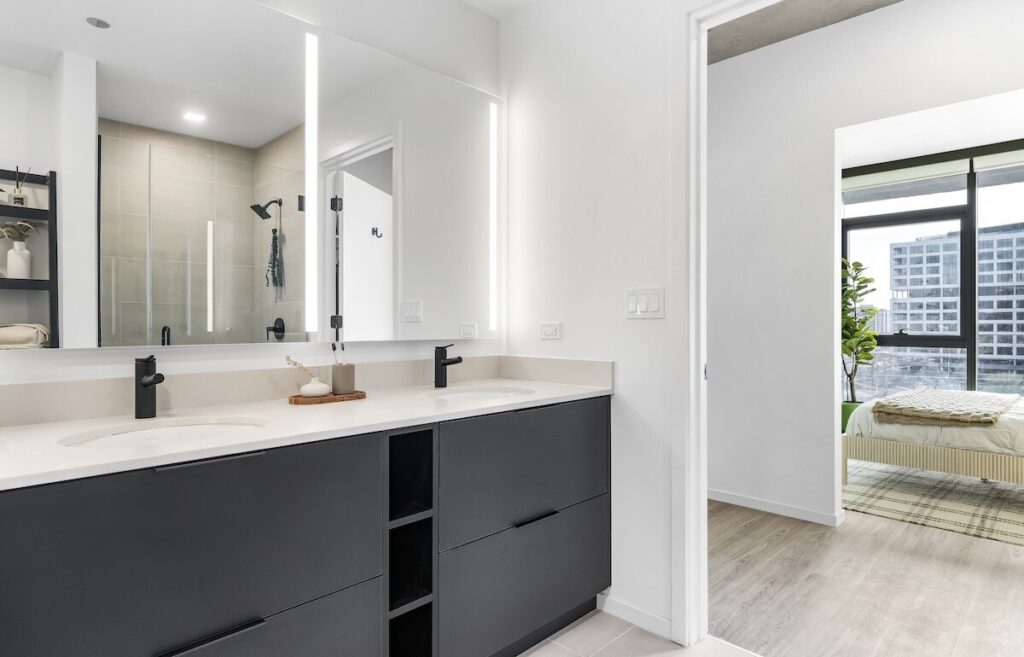 Luxury apartments at The Elizabeth by Sterling Bay. vanity with black cabinets and a large illuminated mirror, with a view through a doorway into a bedroom with a bed and floor-to-ceiling windows.