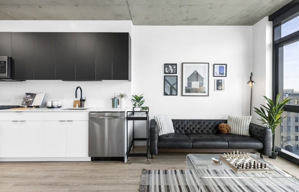 Luxury apartments at The Elizabeth by Sterling Bay. A modern apartment living space featuring a kitchenette with black and white. cabinetry, a black tufted leather sofa, and a glass coffee table with a chessboard. Luxury Apartments for leasing at The Elizabeth