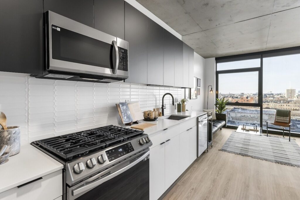 Luxury apartments at The Elizabeth by Sterling Bay. Modern kitchen with matte black upper cabinets, white lower cabinets, and stainless steel appliances, opening into a living area with floor-to-ceiling windows.