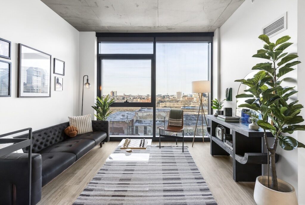 Luxury apartments at The Elizabeth by Sterling Bay. Modern living room with a black tufted sofa, a glass coffee table with a chess set, a chair, and a large window overlooking a city skyline.