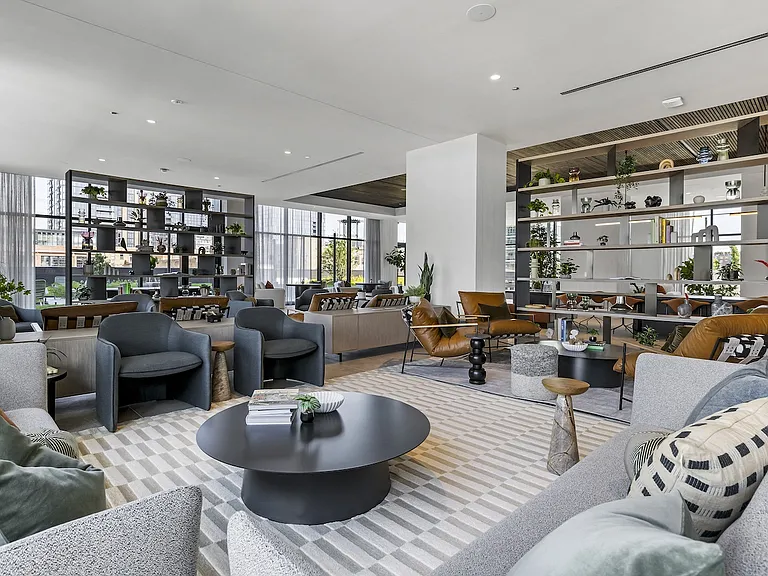 Luxury apartments at The Elizabeth by Sterling Bay. A spacious, modern lounge with various armchairs and sofas arranged around round coffee tables, separated by tall open shelving units.
