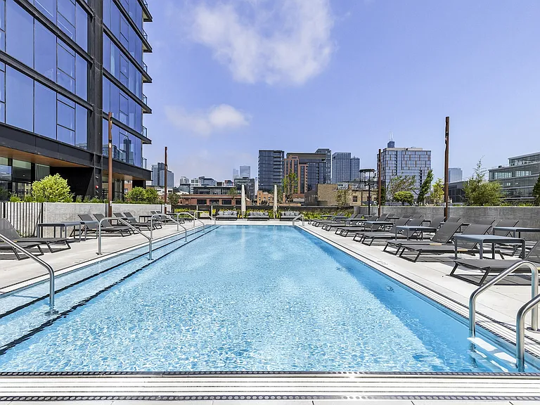 Rectangular outdoor swimming pool lined with lounge chairs, next to a modern high-rise building with a city skyline in the background.