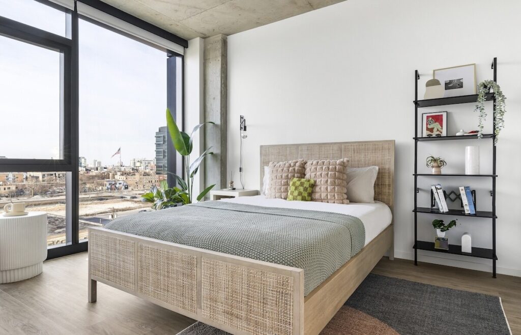 Luxury apartments at The Elizabeth by Sterling Bay. Luxury apartments at The Elizabeth by Sterling Bay. Modern bedroom featuring a cane bed frame with a green throw, floor-to-ceiling windows with a city view, and a black metal shelving unit.
