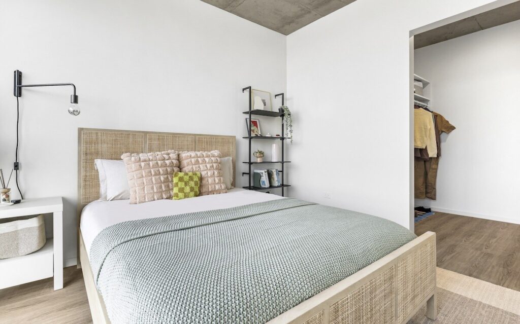 Luxury apartments at The Elizabeth by Sterling Bay. A bedroom with a rattan-frame bed, light green knit throw blanket, and a metal shelving unit next to an open closet.